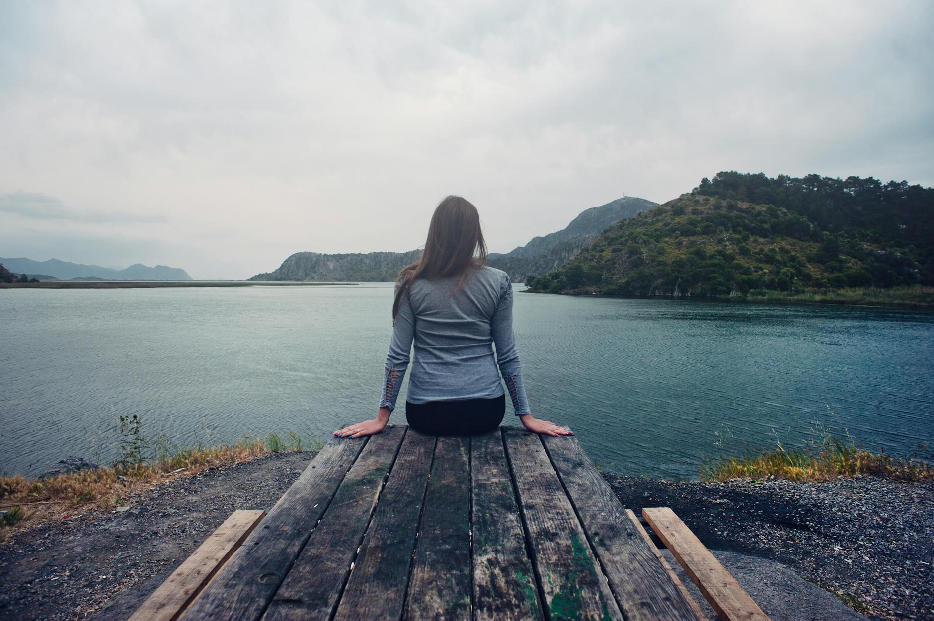 Woman Wearing Gray Long-sleeved Shirt and Black Black Bottoms Outfit Sitting on Gray Wooden Picnic Table Facing Towards Calm Body of Water at Daytime