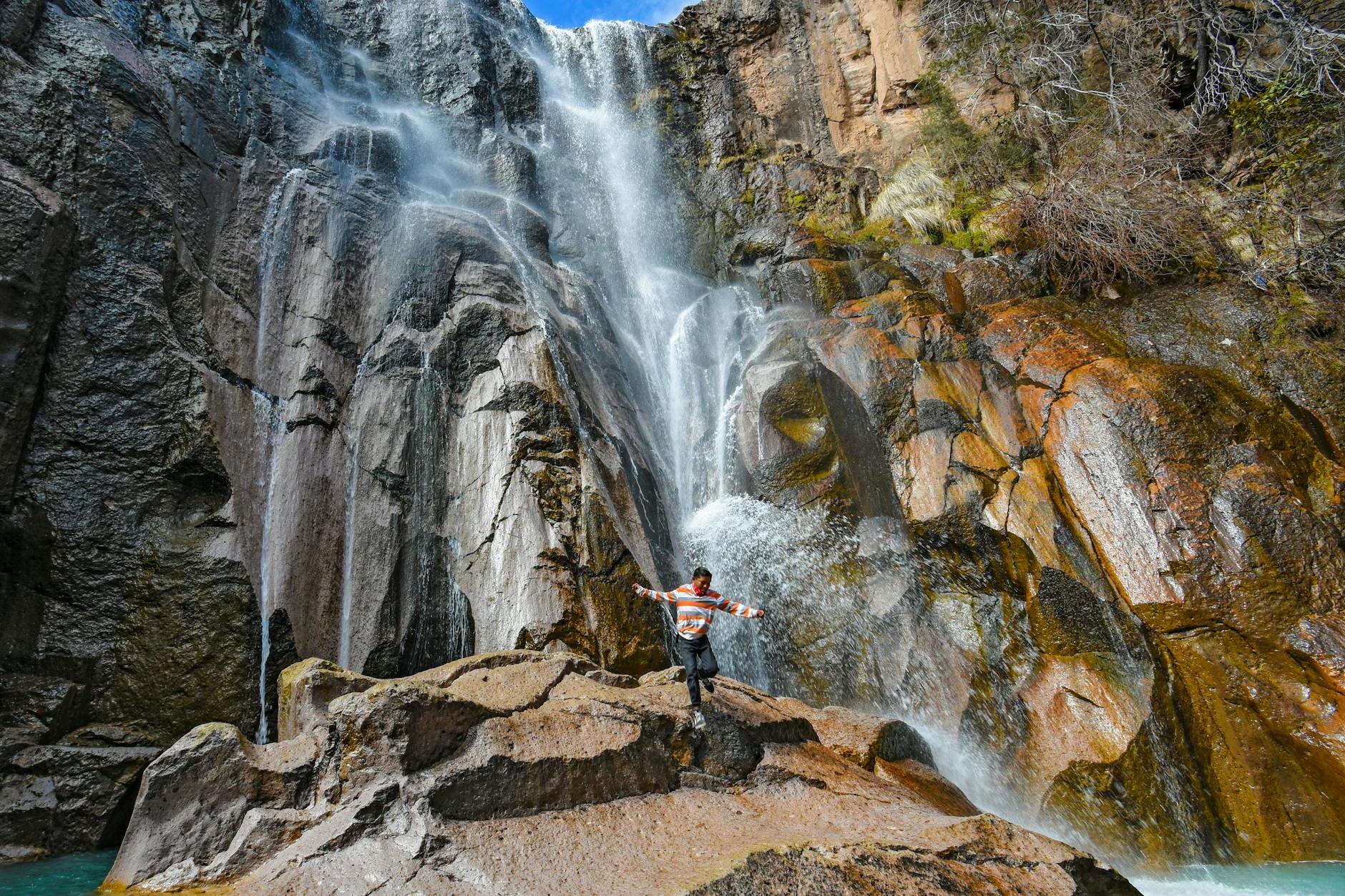 Man in Orange Long Sleeve Shirt Standing on Rock Formation Near Waterfalls