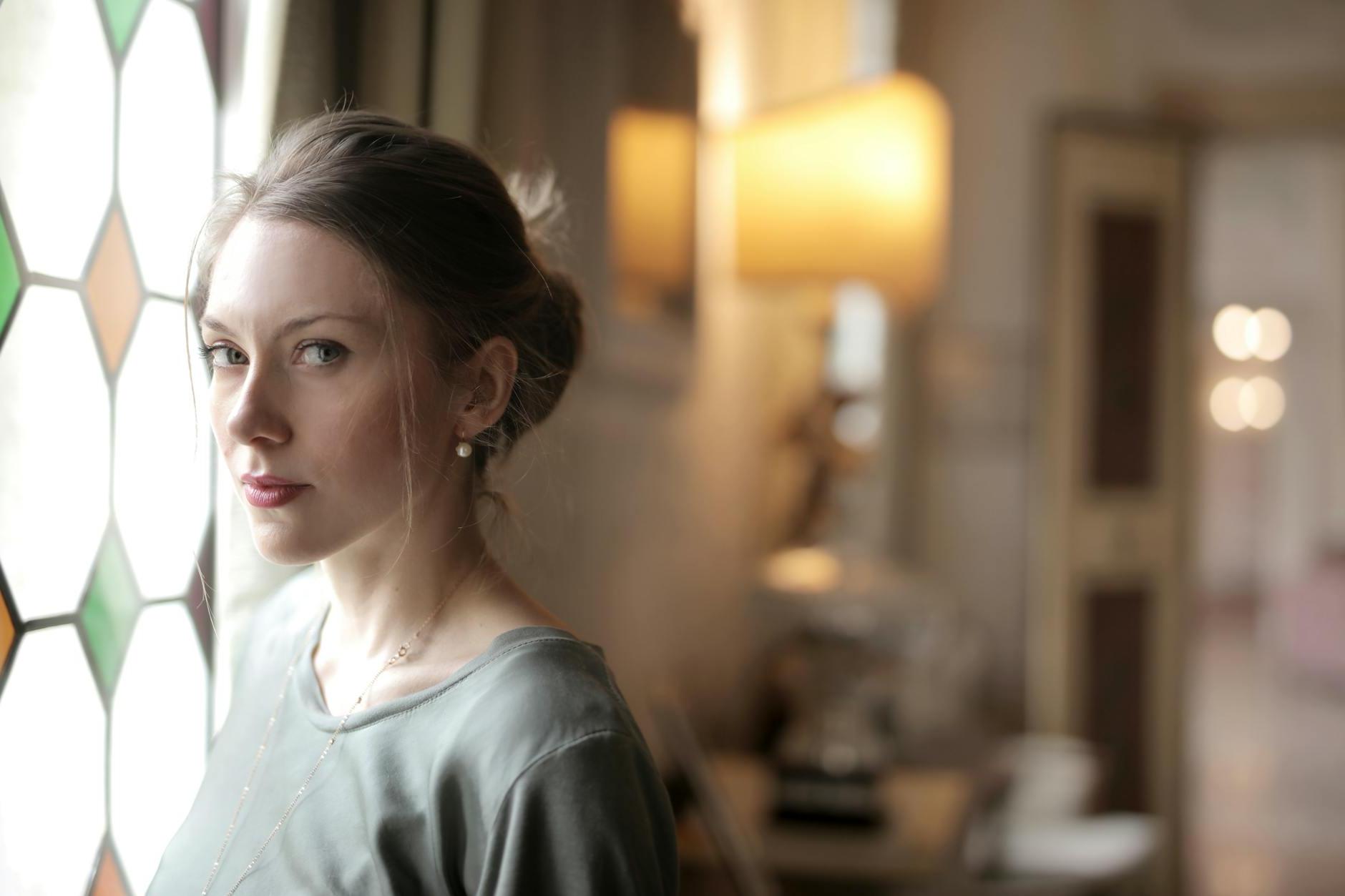 Pretty casual serious lady with hairstyle intently looking at camera while standing near stained glass window in bright luxury room of old mansion