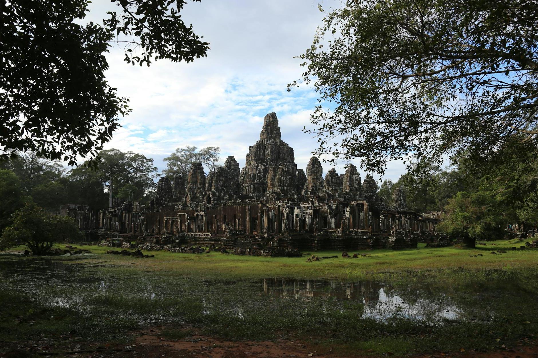 Scenery view of aged church complex exterior on green field near rippled pond and growing trees under cloudy sky in Cambodia