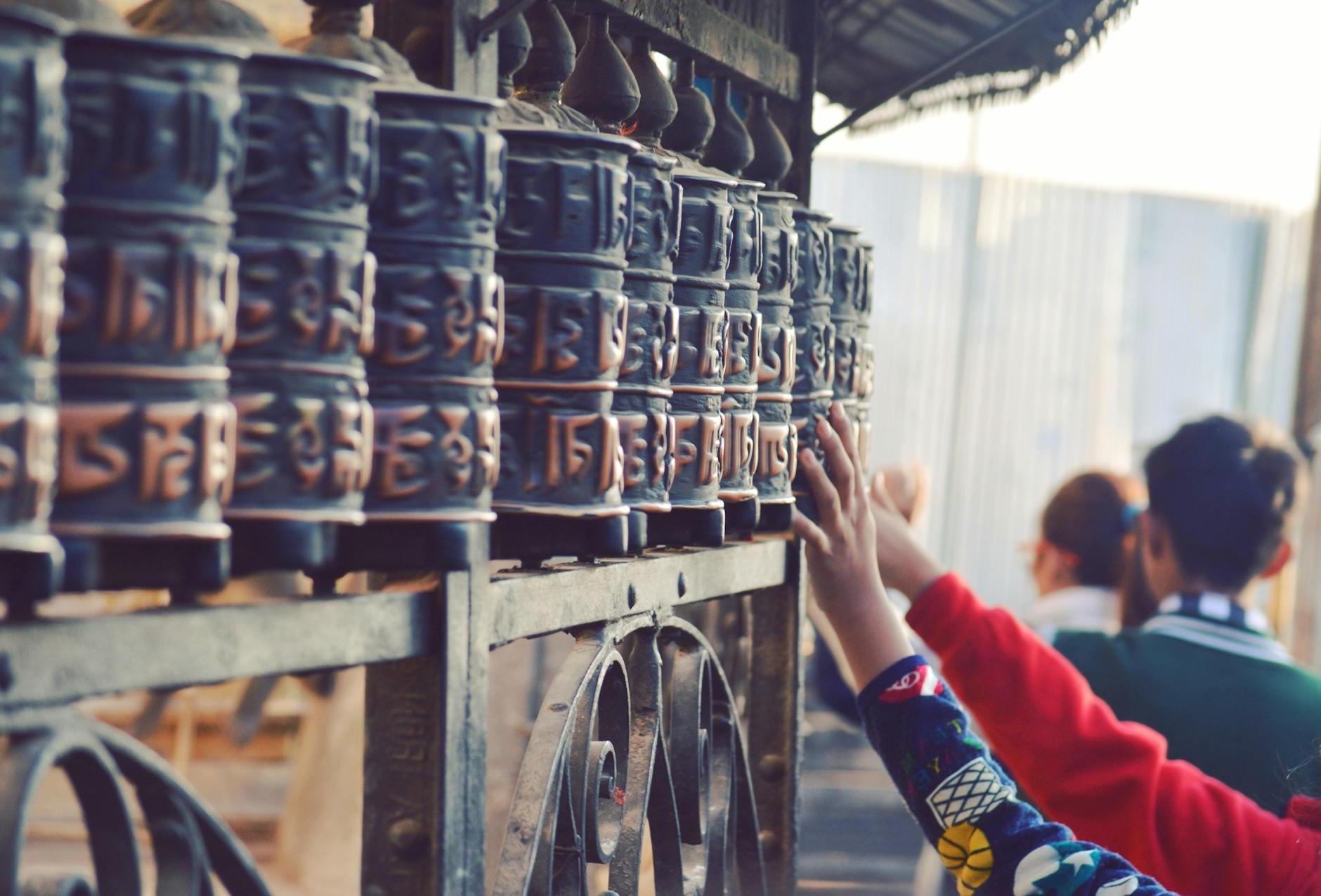 Back view of anonymous tourists touching old metal Prayer wheels with hieroglyphs above ornamental fence near old Buddhist temple in Swayambhunath