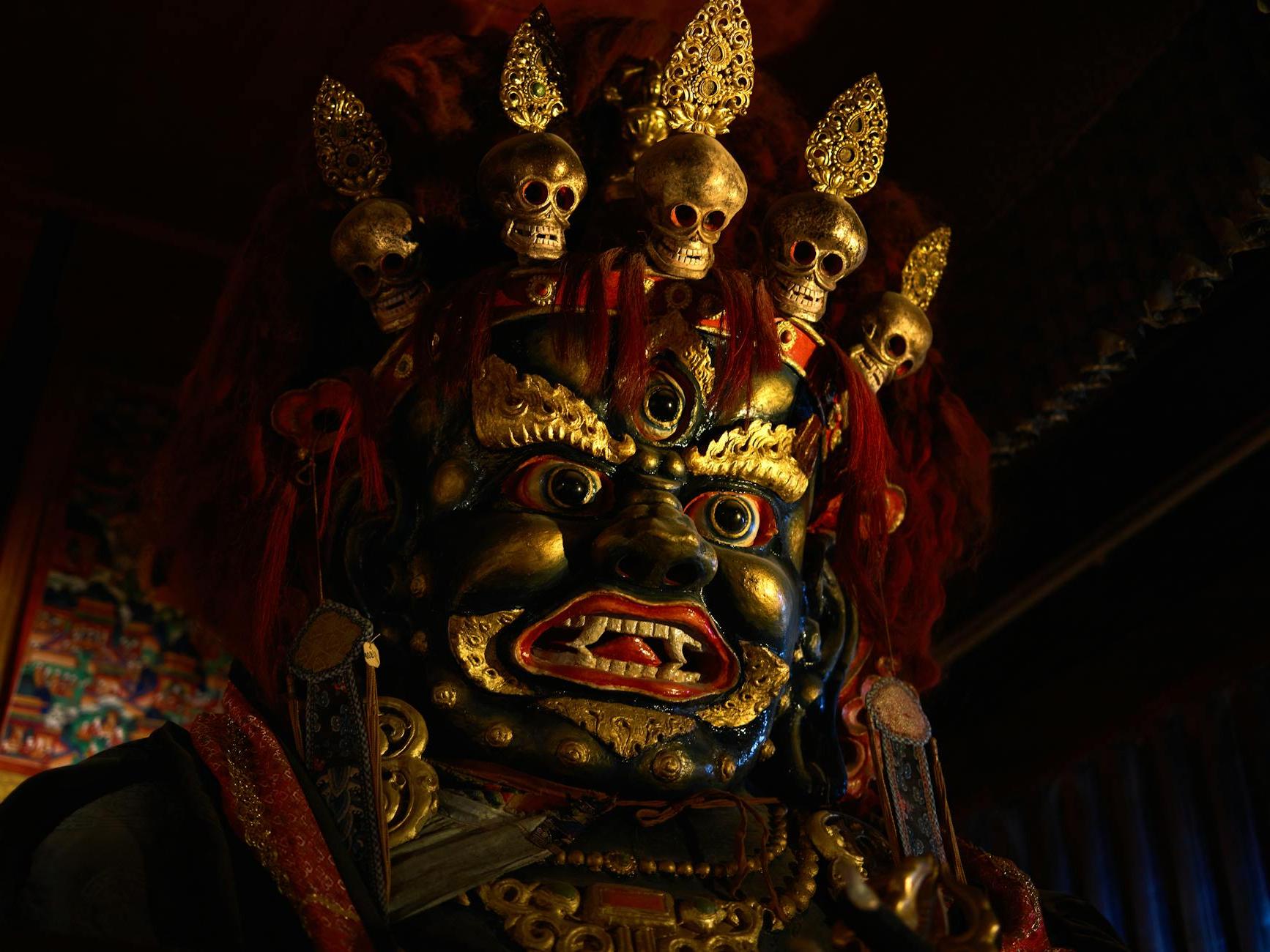 From below of Tsam mystery mask with gilded human face and third eye and pig nose and fangs among teeth and golden circles around face and full head of red hair decorated with red hairband adorned with golden skulls