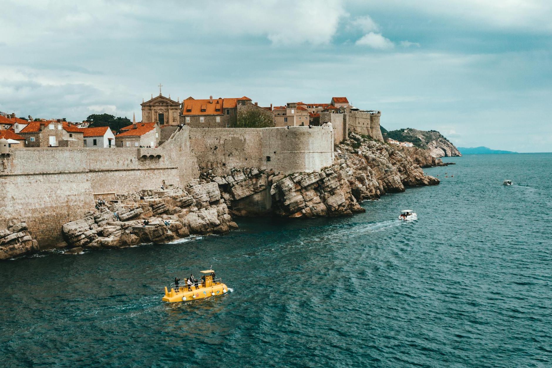 Modern boats floating on rippling sea near rocky coast of old town of Dubrovnik with historical buildings and ancient city walls