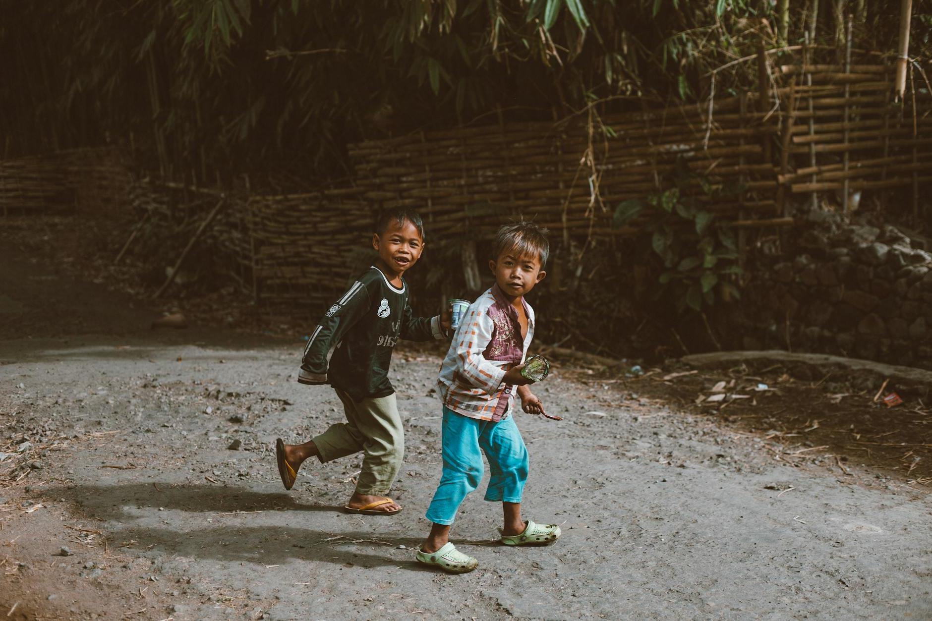 Full body of little ethnic children walking on dirty path near green plants in village on sunny day