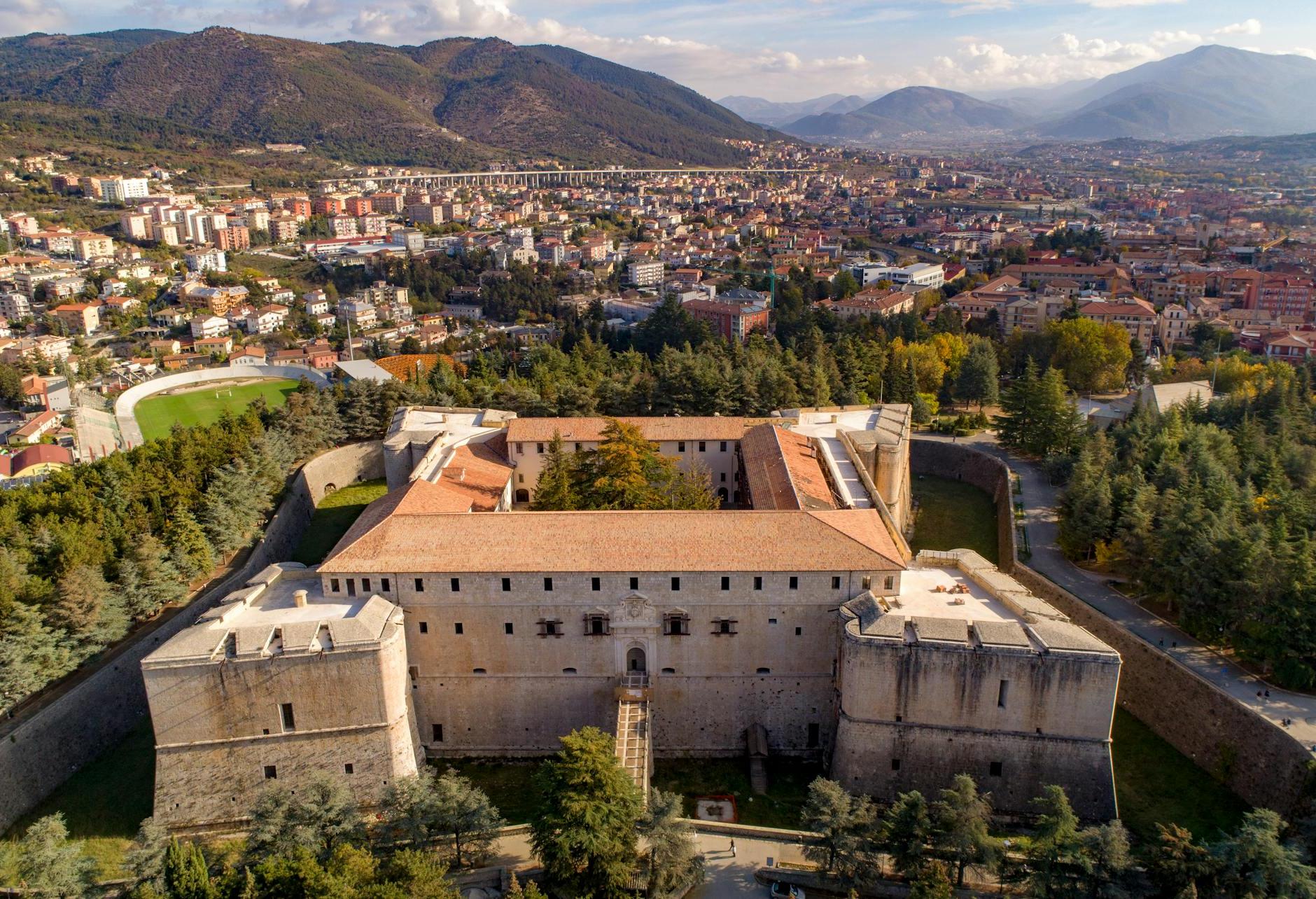 Picturesque view of ancient stone castle with green plants and stone towers surrounded by medieval brick walls and green trees located near town and mountains