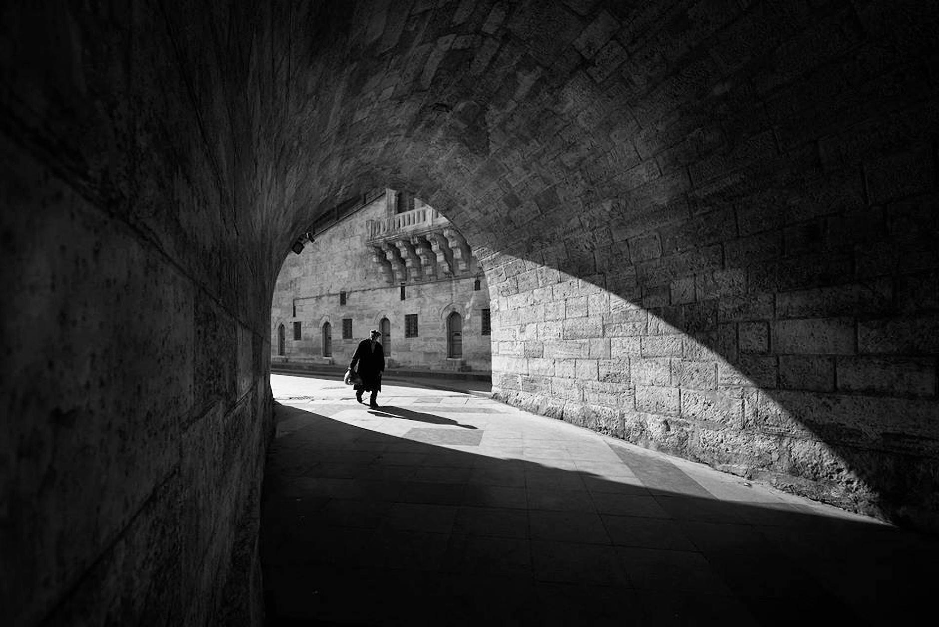 Black and white anonymous male walking along old stone tunnel near aged brick building