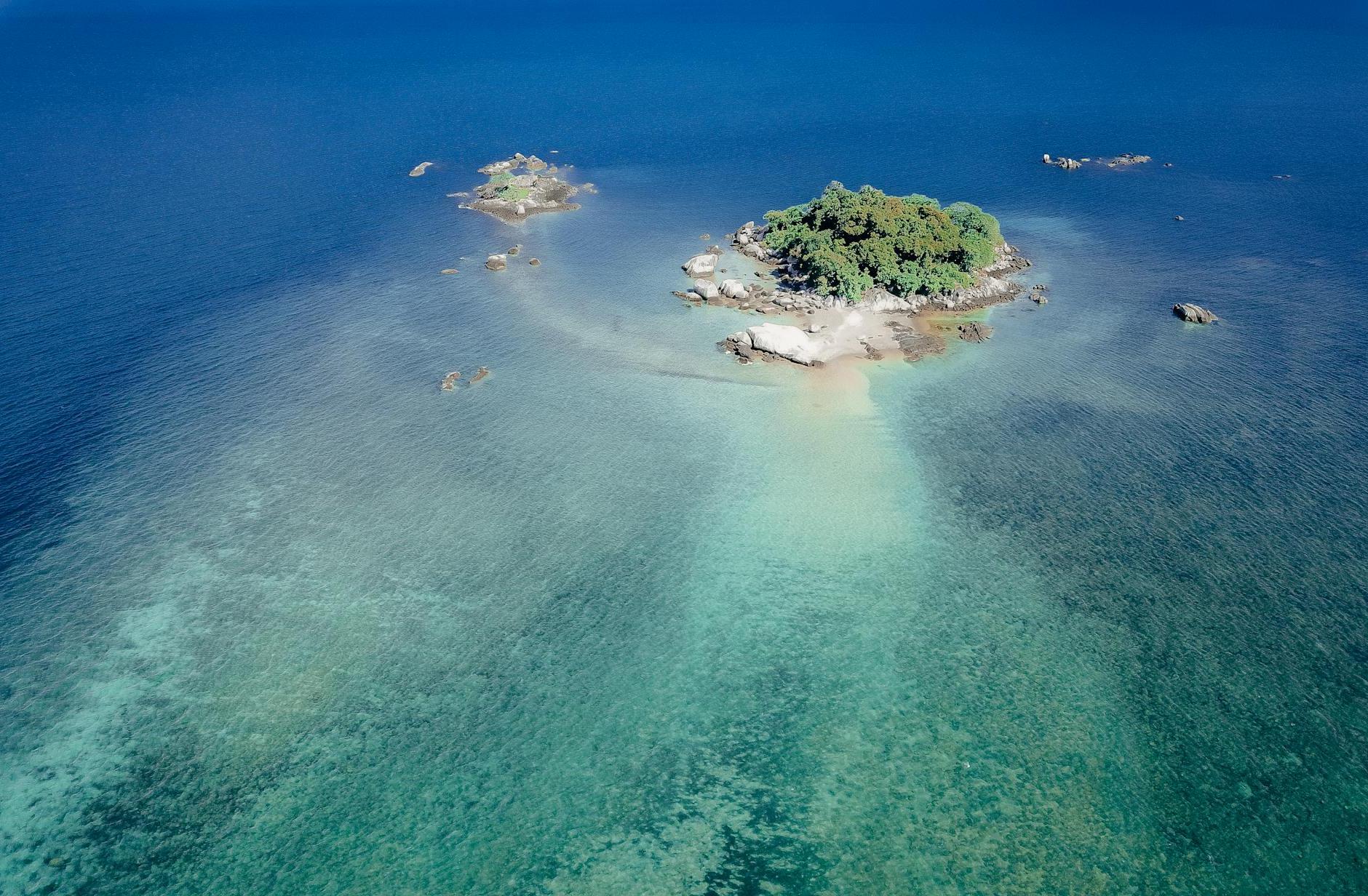 Drone view of turquoise shallow sea with small cay covered with lush tropical greenery in sunlight