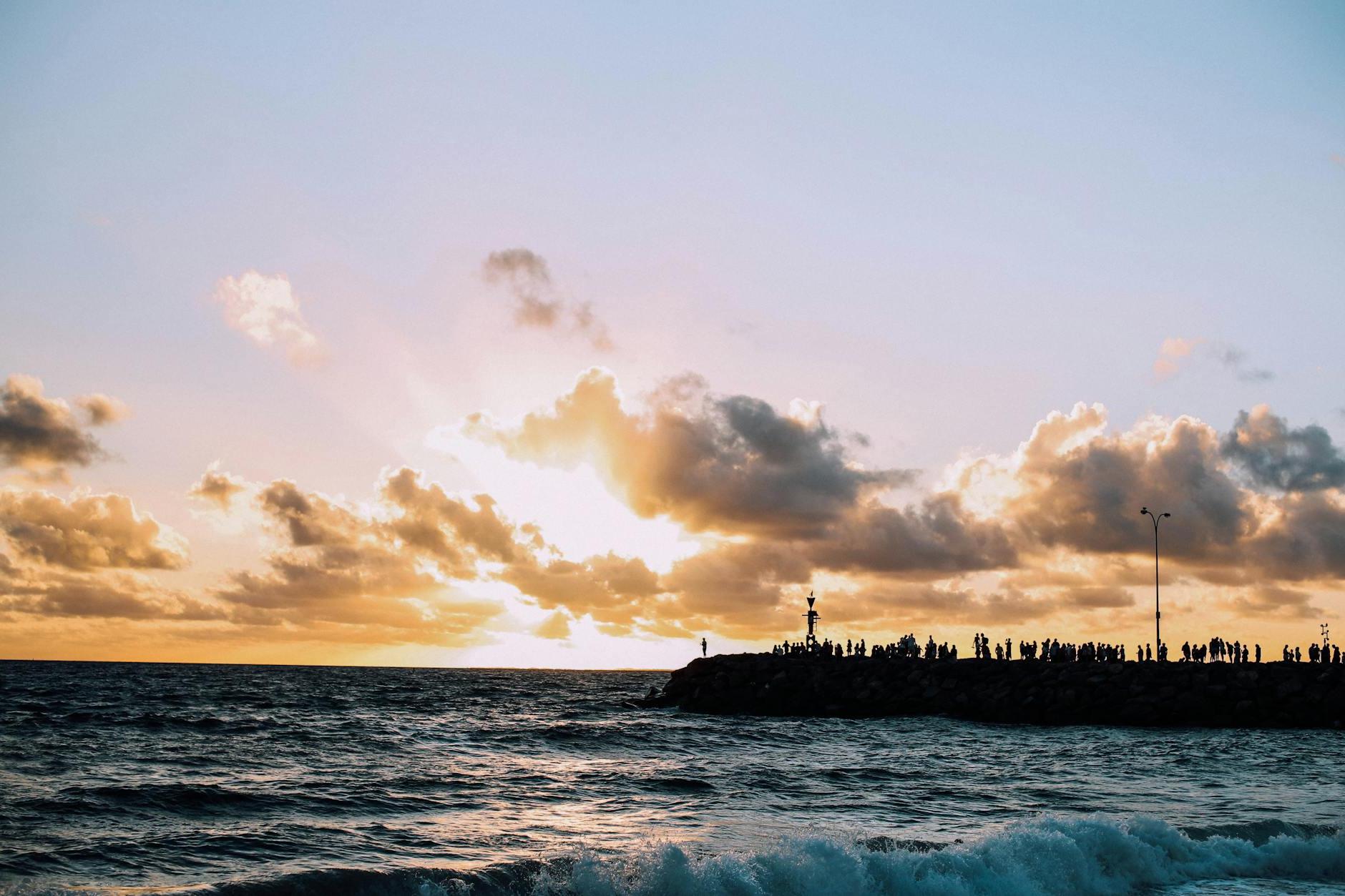 Silhouettes of people standing on embankment of sea with splashing waves under clouds at sunset