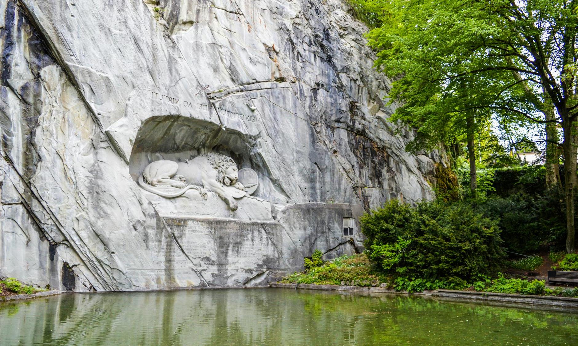 Sculpture of a mortally wounded lion carved into sheer rock ledge near pond located in Switzerland