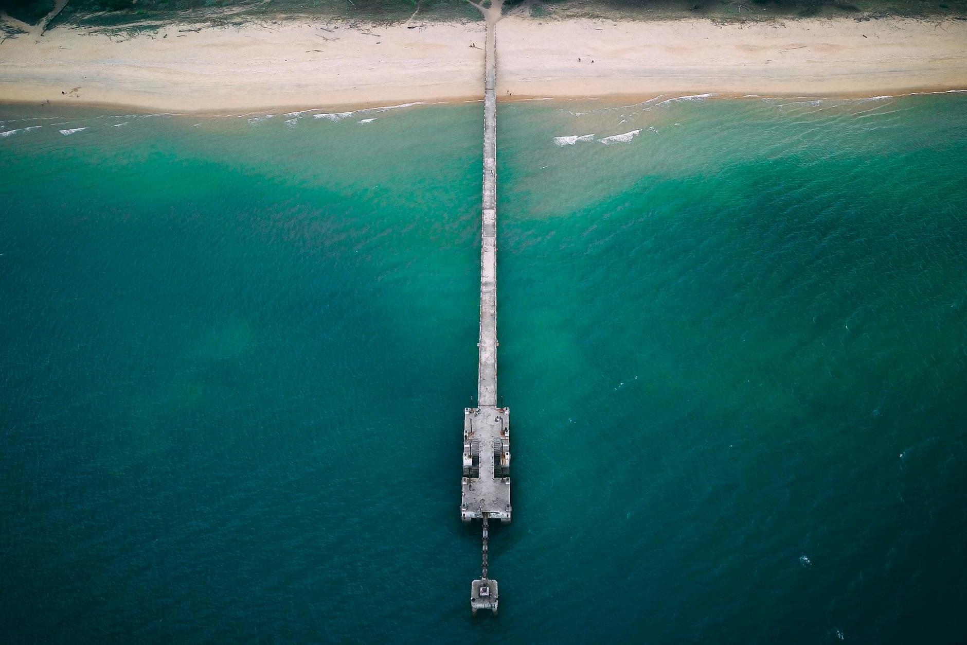 Drone view of long pier near sandy shore washed by turquoise water of blue endless ocean