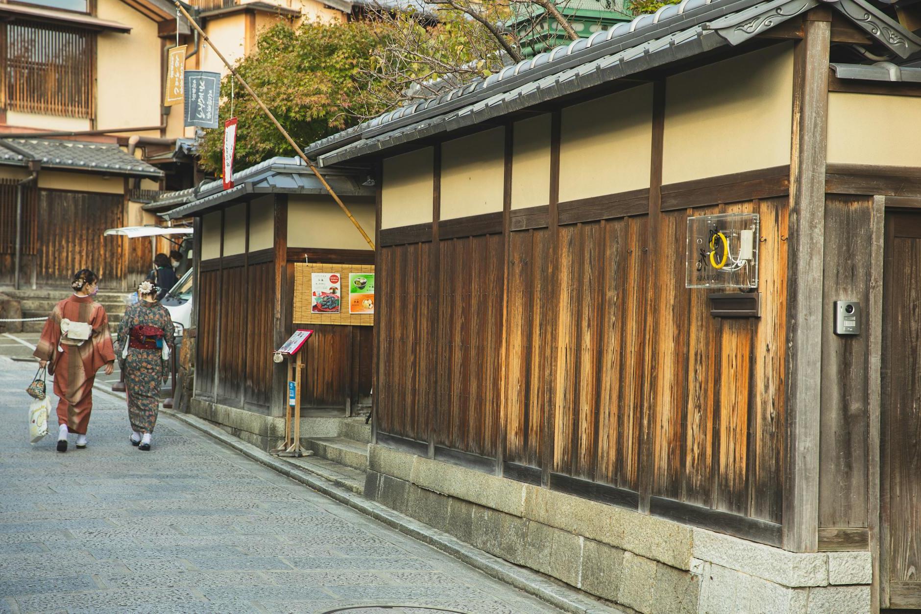 Back view of unrecognizable local Japanese ladies in traditional kimonos walking on paved street near typical aged wooden houses in Kyoto