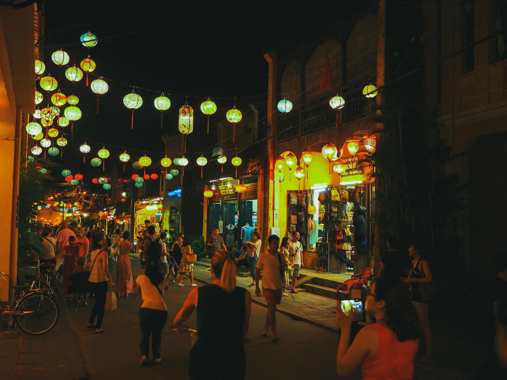 Crowd of unrecognizable diverse tourists walking on narrow pedestrian street with aged houses and cafes in city old district decorated with Asian paper lanterns at night