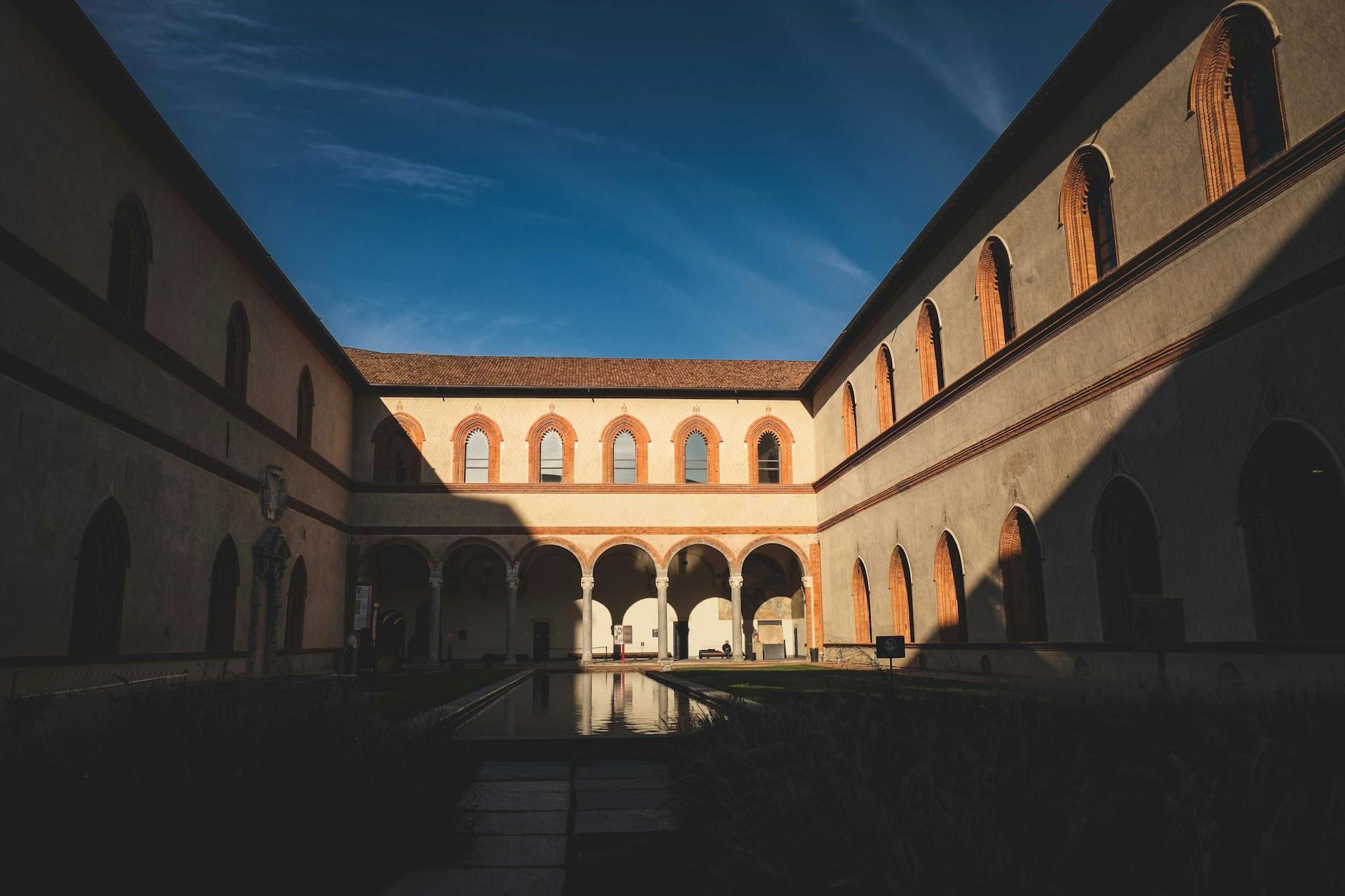 Garden in Ducal Courtyard of historic Castello Sforzesco with arched windows and passage against bright blue sky in Milan