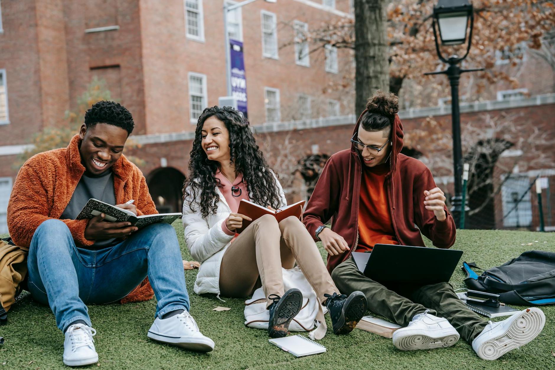 Full body of happy diverse students with notebooks and laptop sitting on grassy lawn on campus of university while studying together