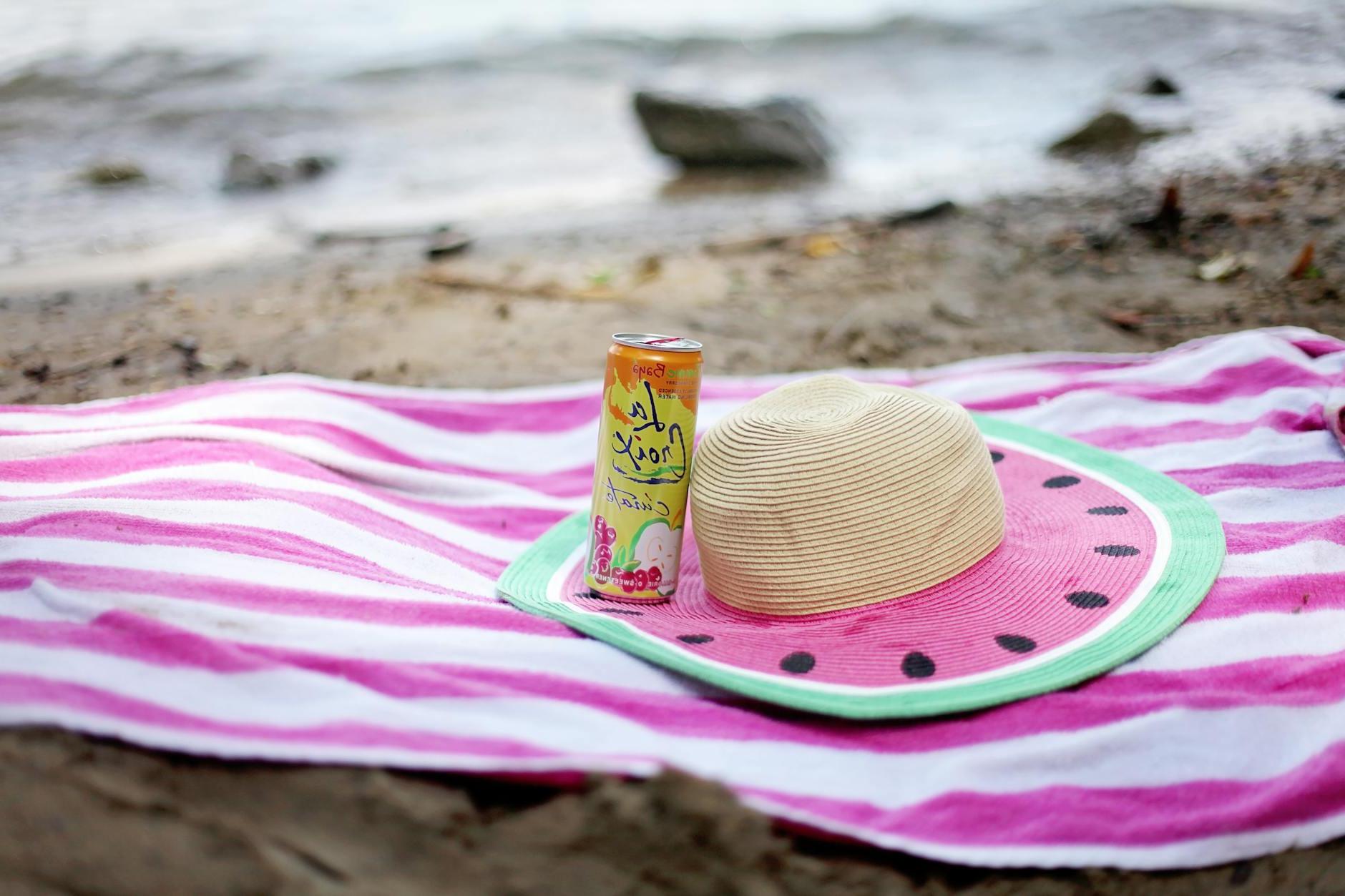 Striped towel spread on sandy beach with straw hat and juice can
