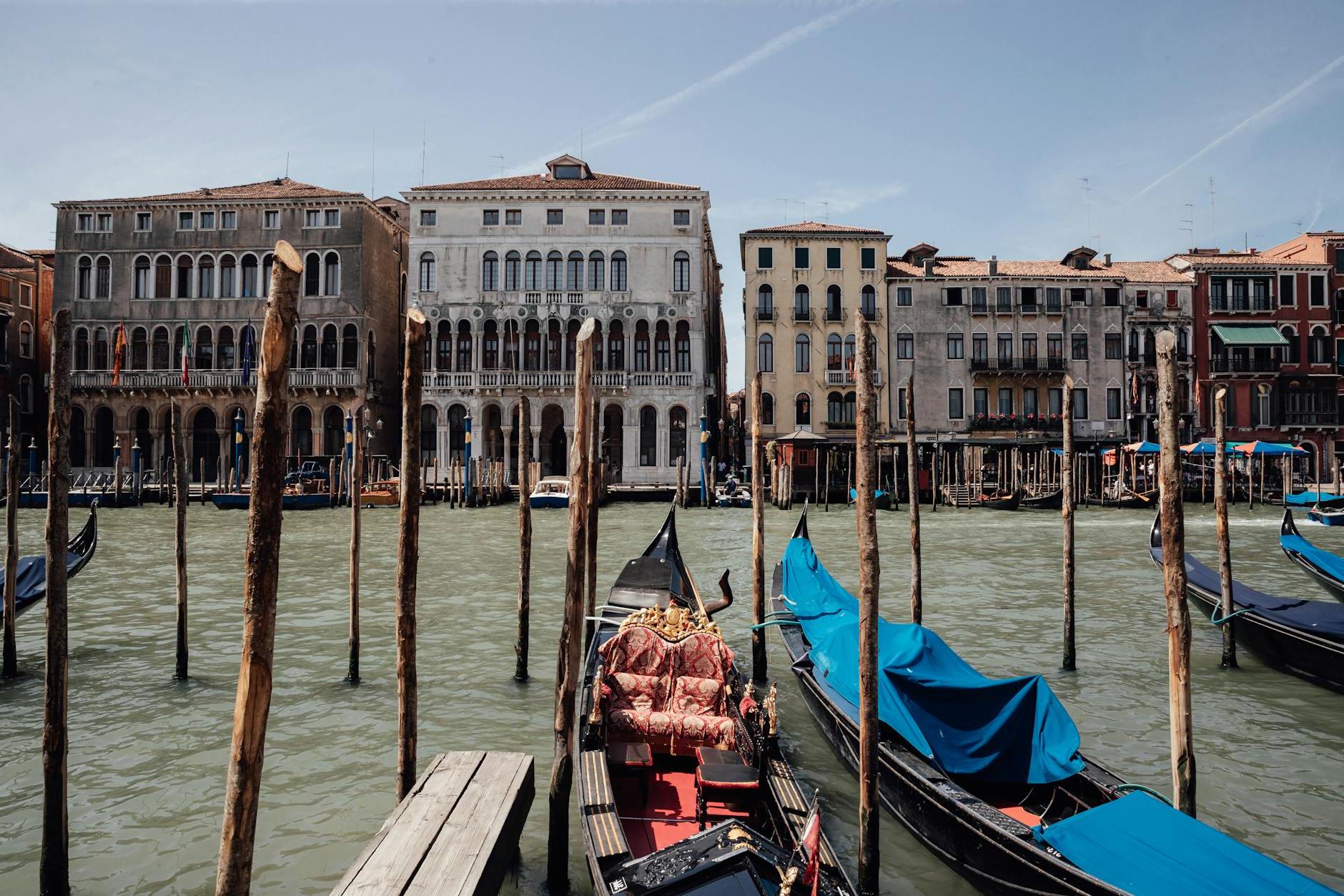 Gondolas moored on Grand Canal with wooden poles against facades of aged shabby residential buildings and blue sky in Venice