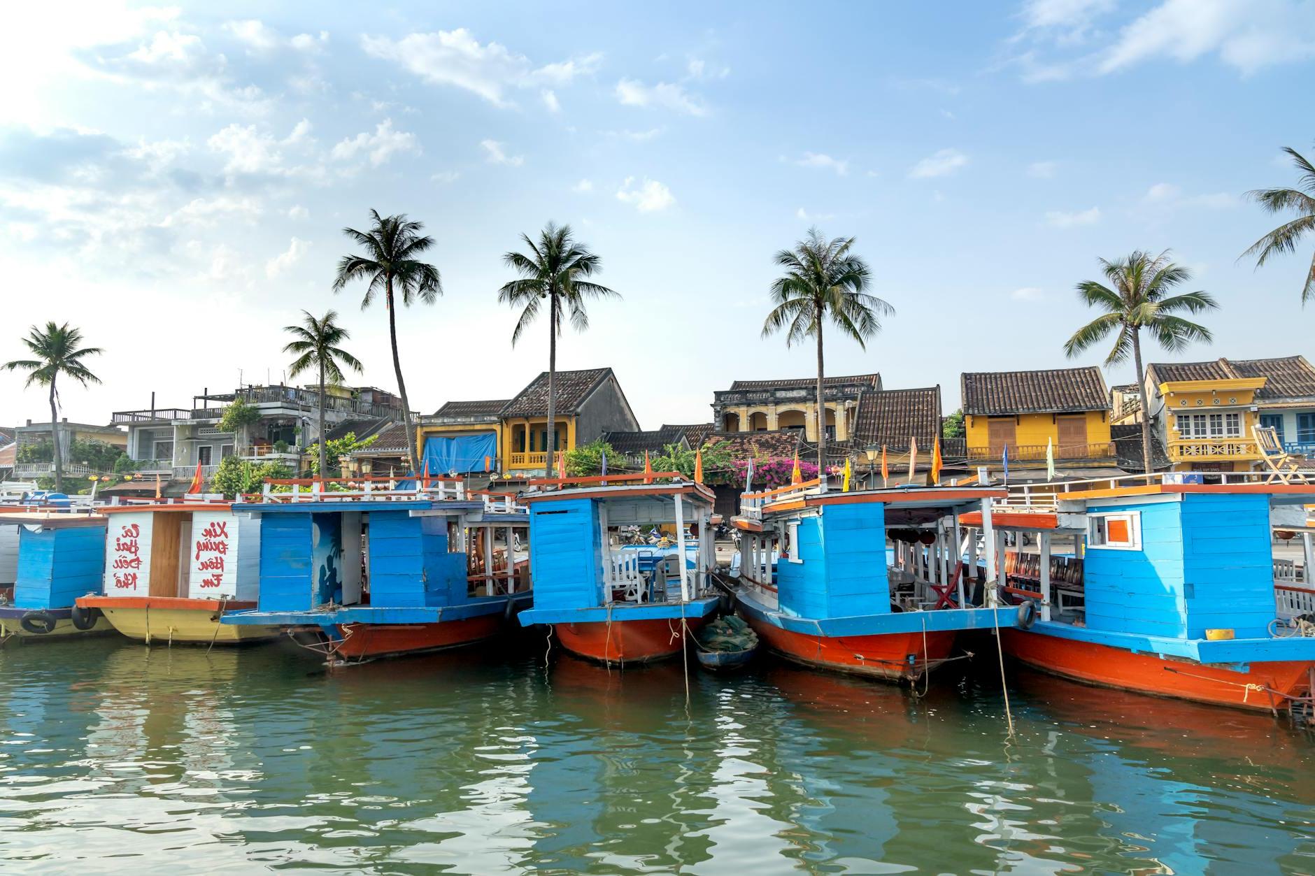 Excursion boats moored on river shore near typical residential cottages on sunny day in Hoi An