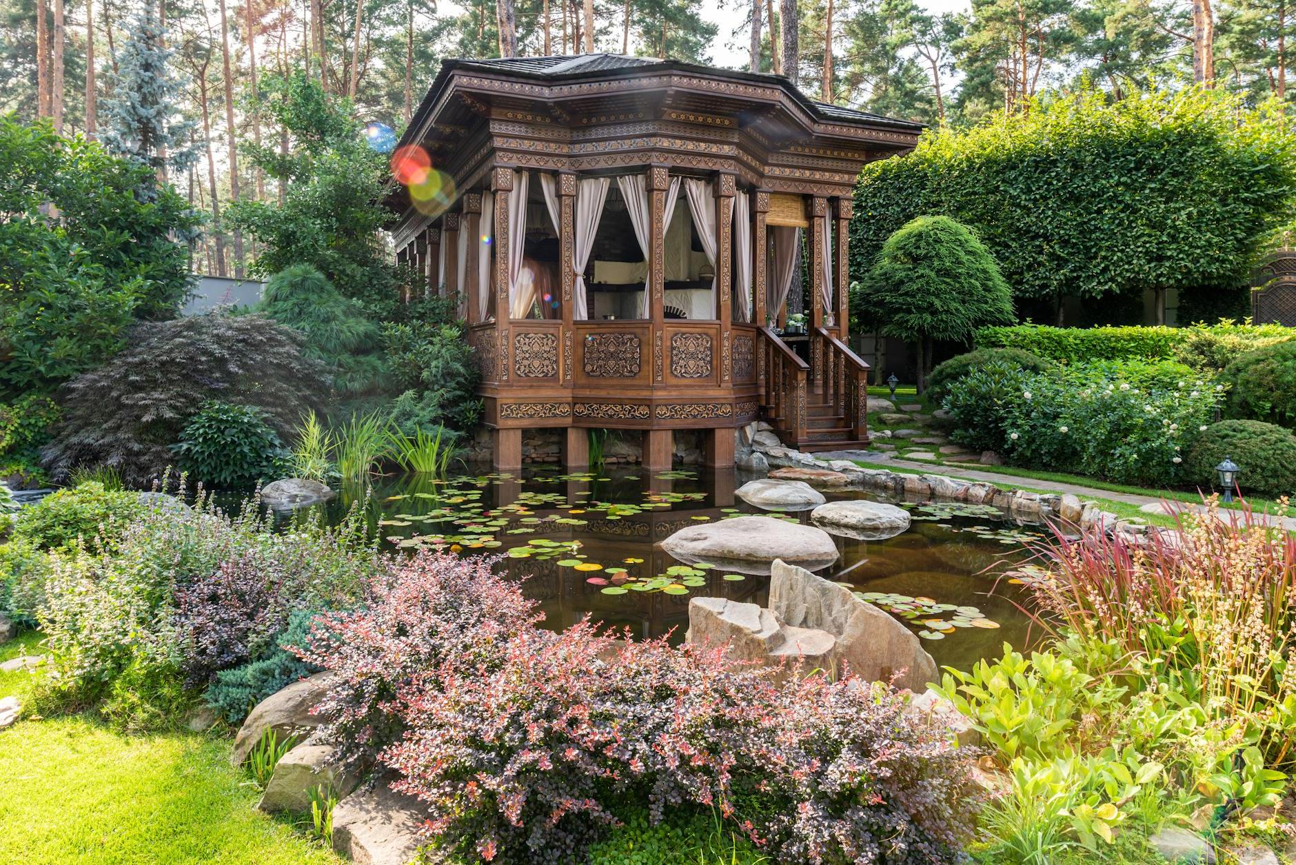 Aged wooden summer house near pond with rocks near green plants and flowers in garden in sunny day