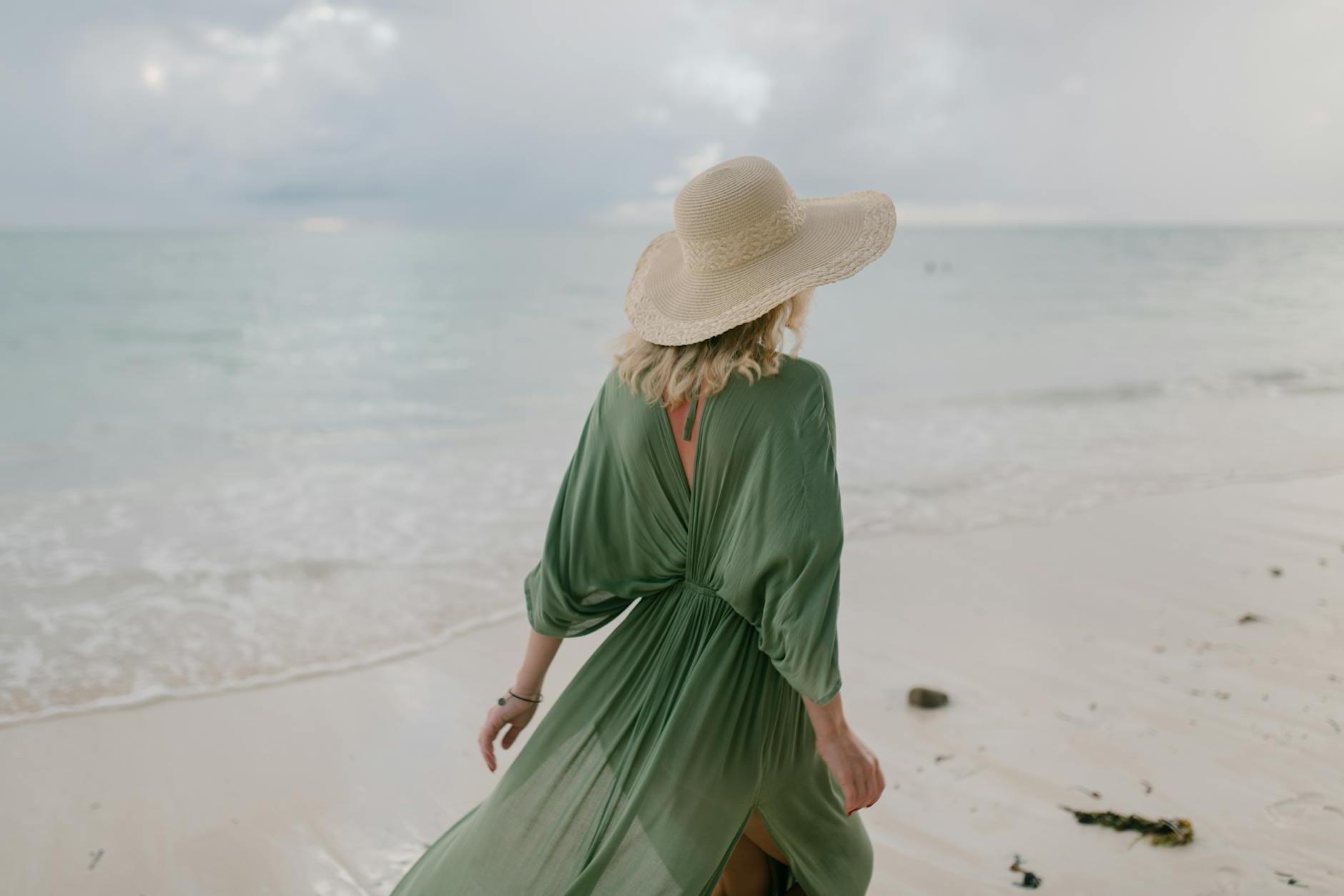 Back view of unrecognizable female tourist wearing straw hat and summer dress standing on seashore in cloudy day