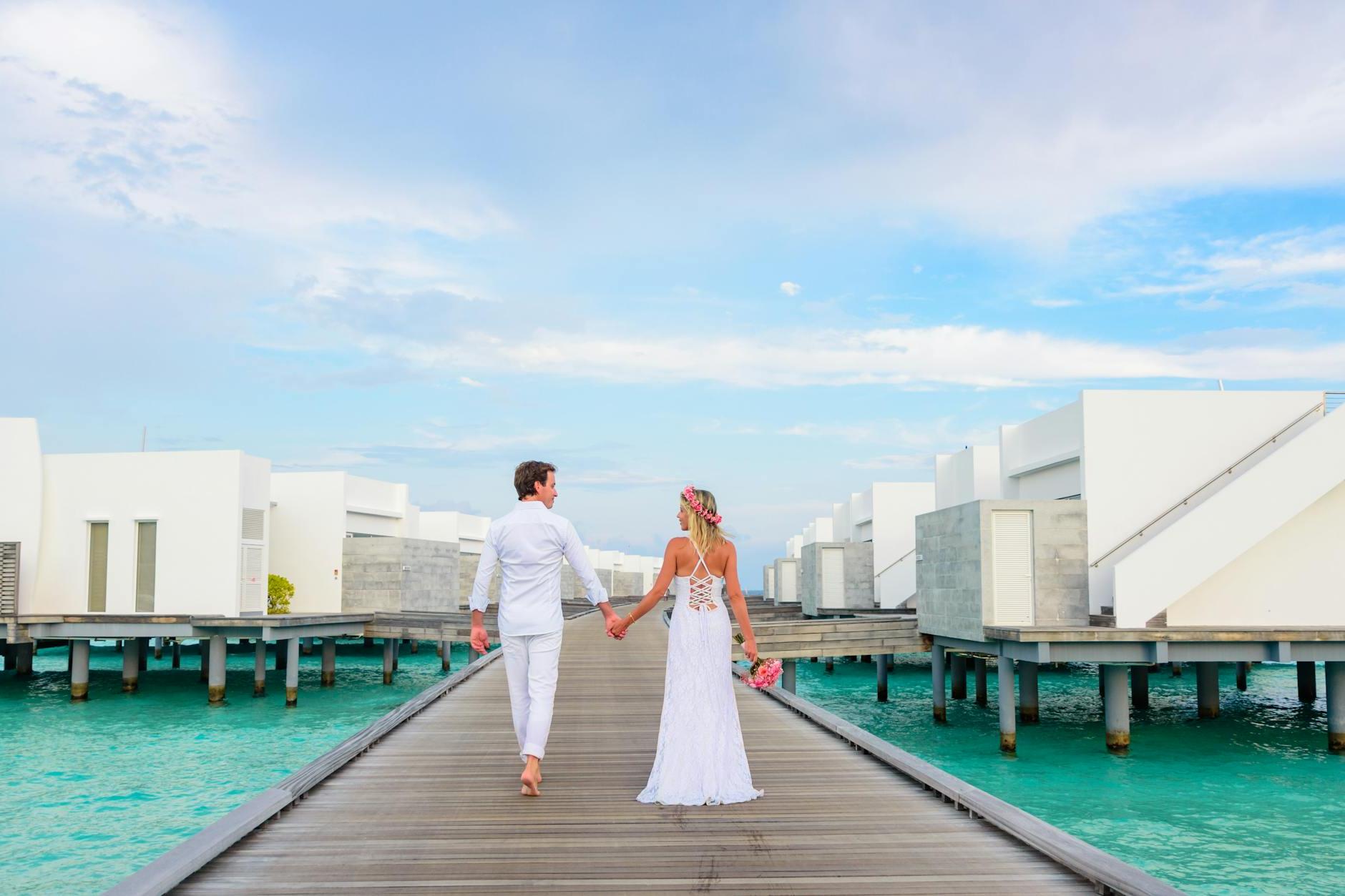 Full body back view of unrecognizable loving couple in white clothes walking on boardwalk above water near buildings while holding hands and looking at each other