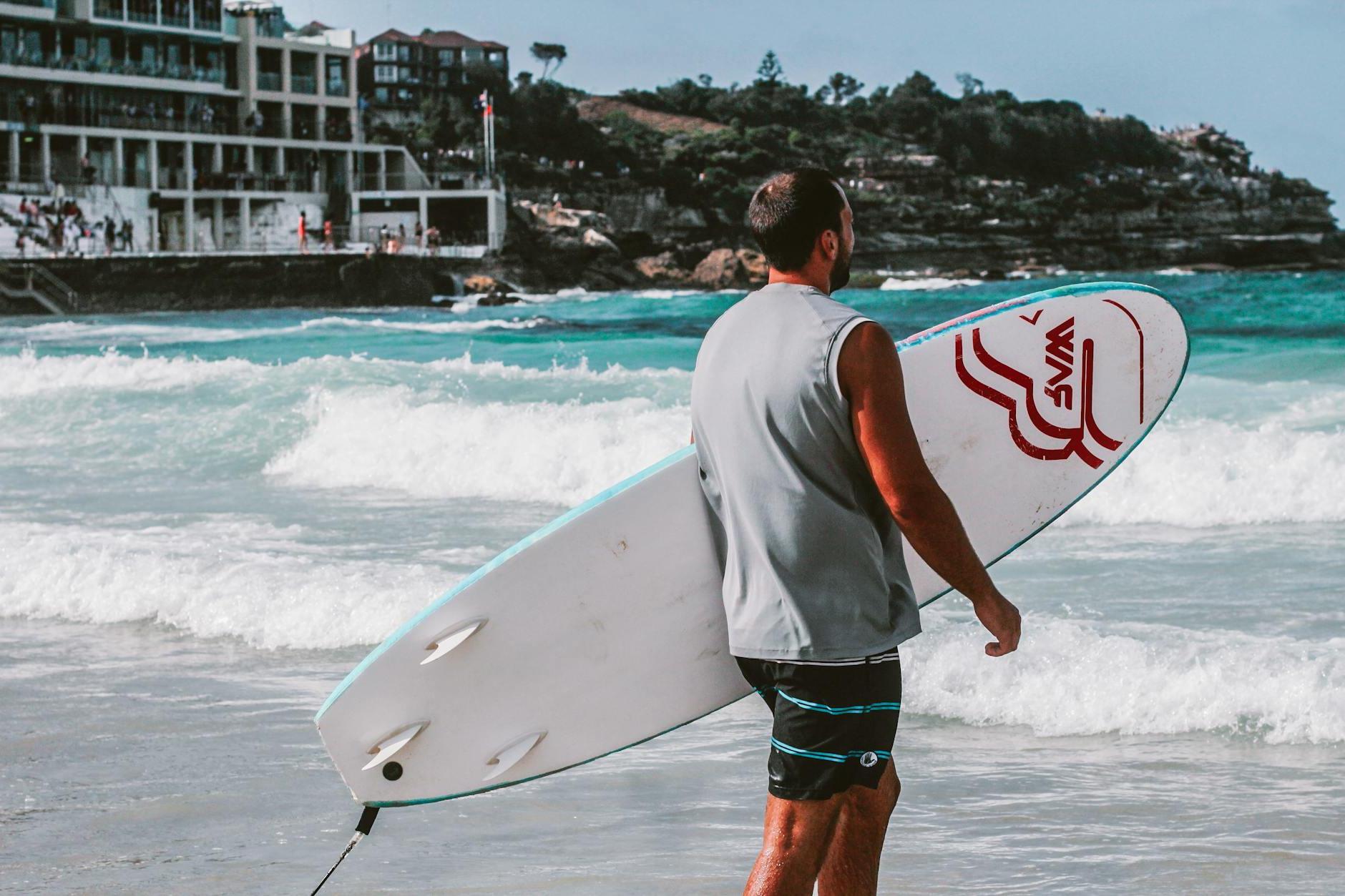 Man Walking Towards Body Of Water Holding A Surfboard